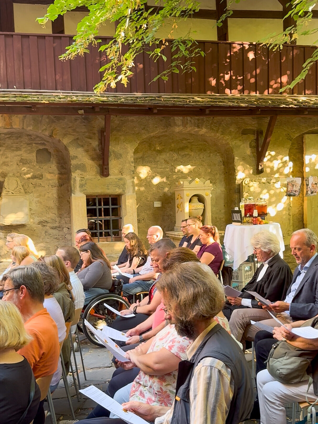 Besucher*innen des Open Air Segnungsgottesdienstes des Christophoruswerks Erfurt sitzen im historischen Innenhof der Michaeliskirche und singen gemeinsam. Sie blicken auf die Notenblätter und werden von der Abendsonne beschienen.